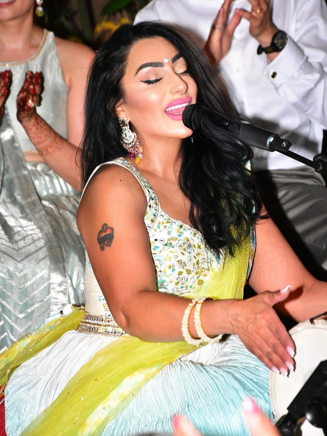 Singer mid-phrase at a Mehndi with a dholki beside her and a guest&rsquo;s henna-covered hands behind
