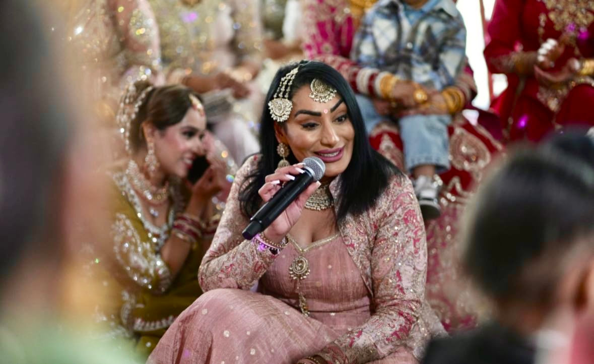 Reena Kaur singing into a microphone at a Ladies Sangeet, with guests seated around her