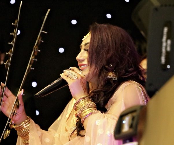 Reena Kaur singing on stage beside a stringed instrument under stage lighting