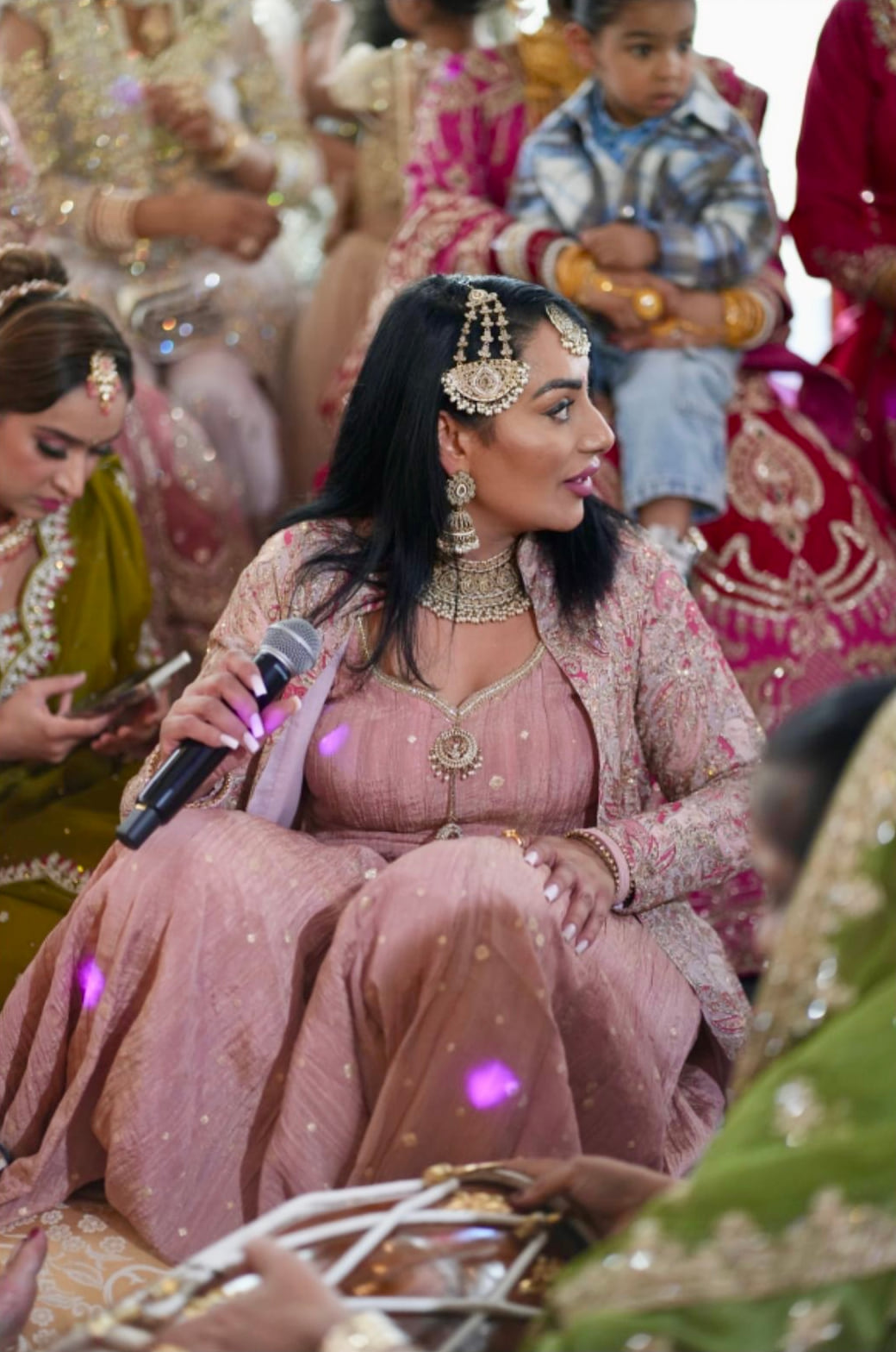 Reena Kaur in a pink embroidered outfit singing at a sangeet, surrounded by seated guests