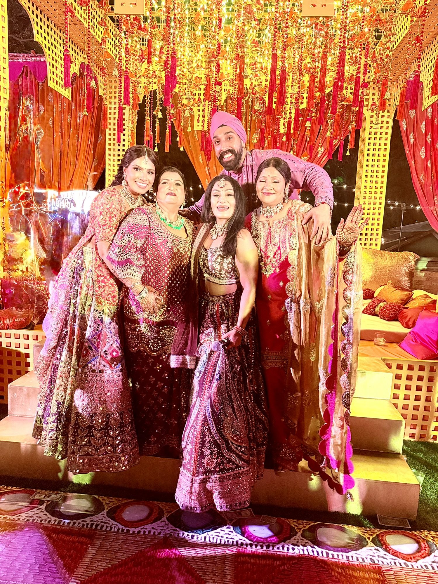 Guests in embroidered lehengas at a Sangeet beneath a gold and red tassel canopy