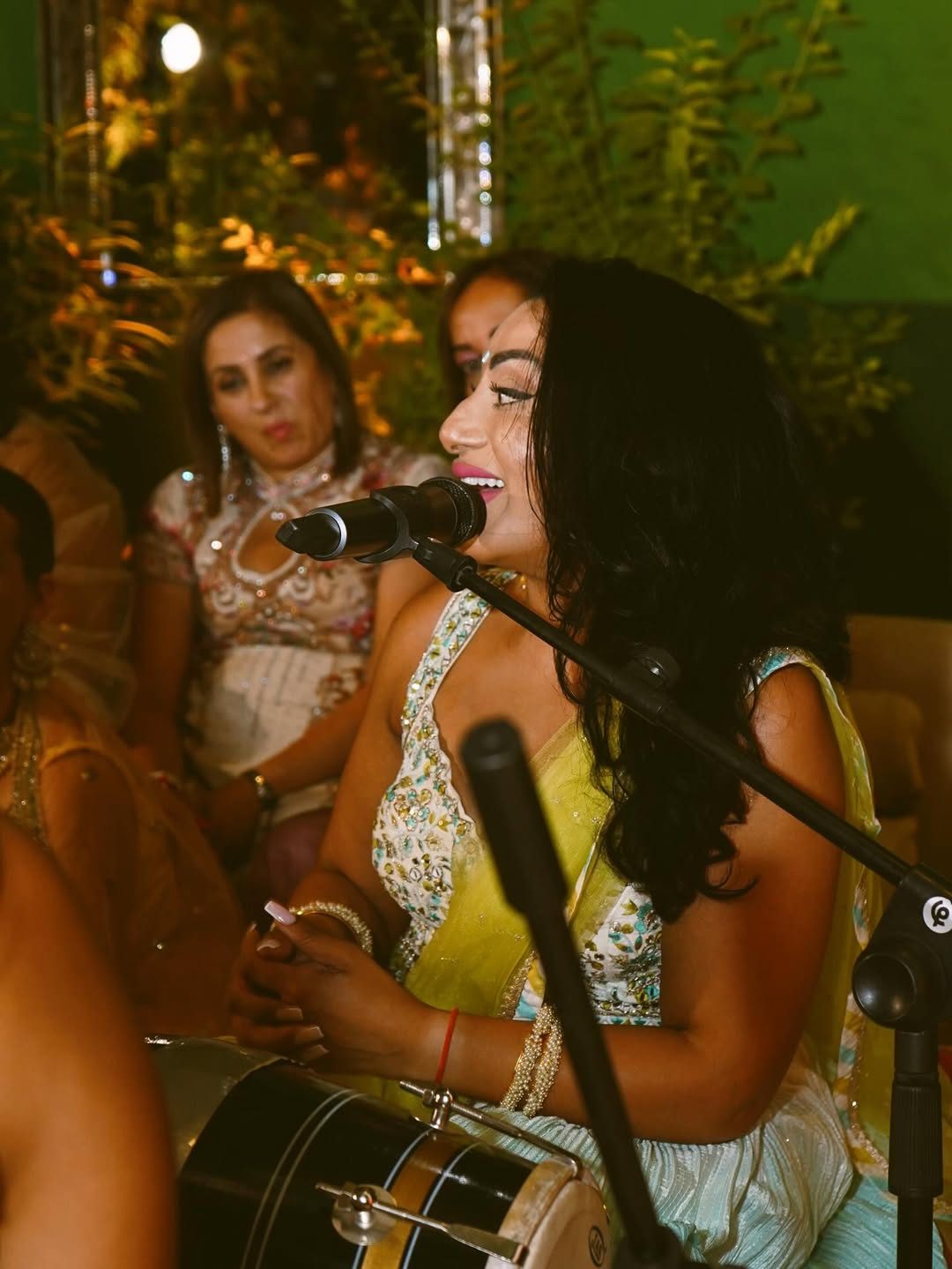 Singer performing at a Sangeet with a dholki, seated guests listening behind her