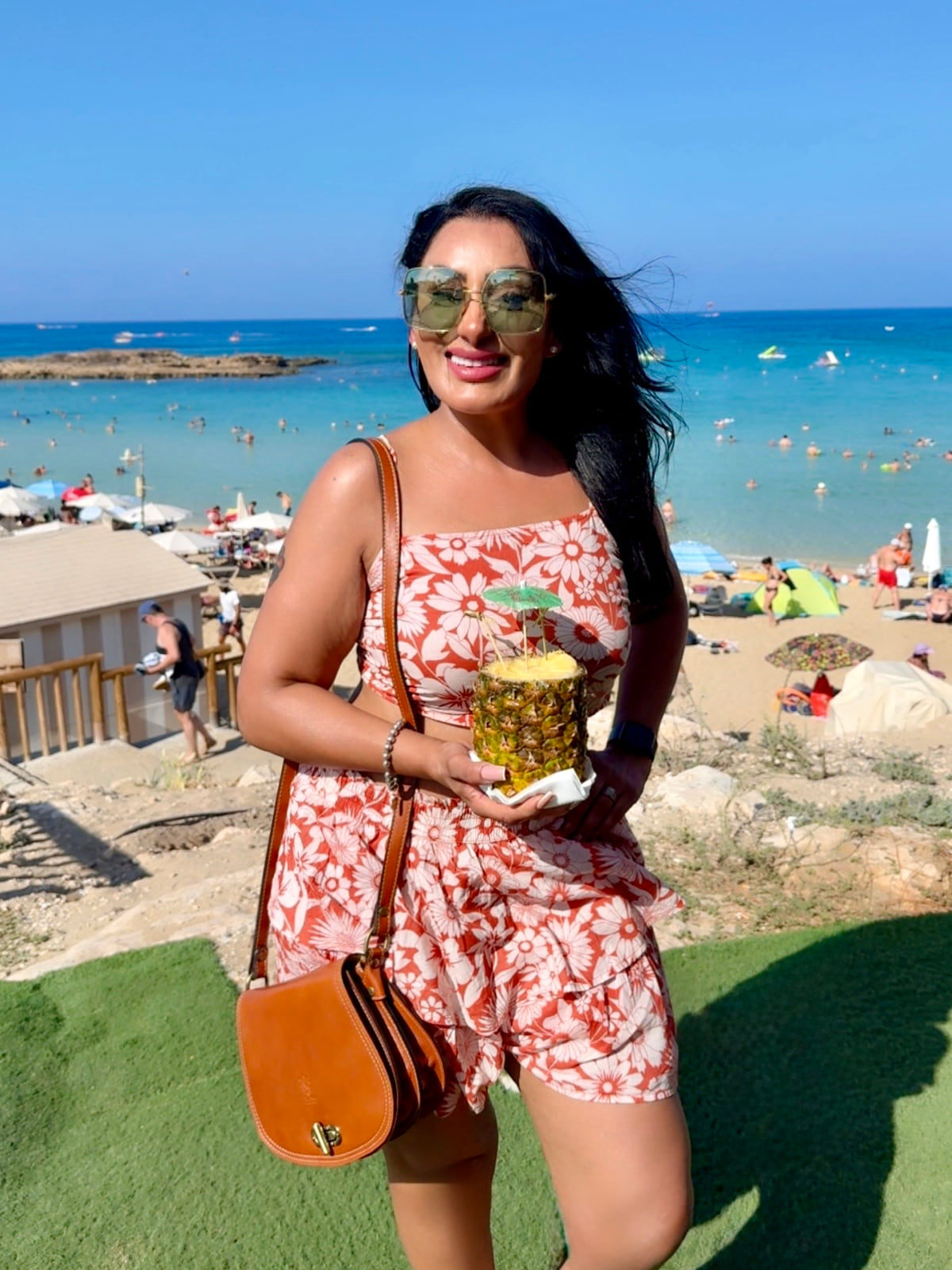 Reena Kaur in a red floral two-piece and sunglasses, holding a pineapple drink above a sunny beach
