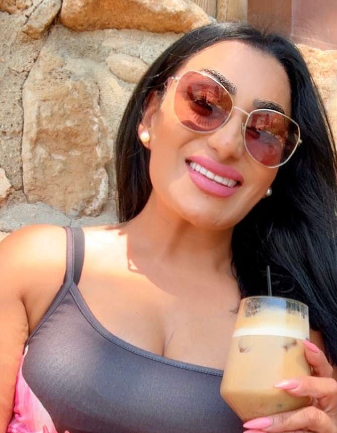 Reena Kaur in sunglasses and a grey vest top, smiling with an iced coffee in the sunshine