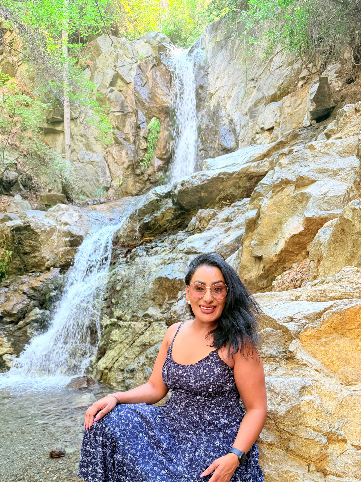 Reena Kaur in a blue floral sundress sitting on the rocks beside a waterfall