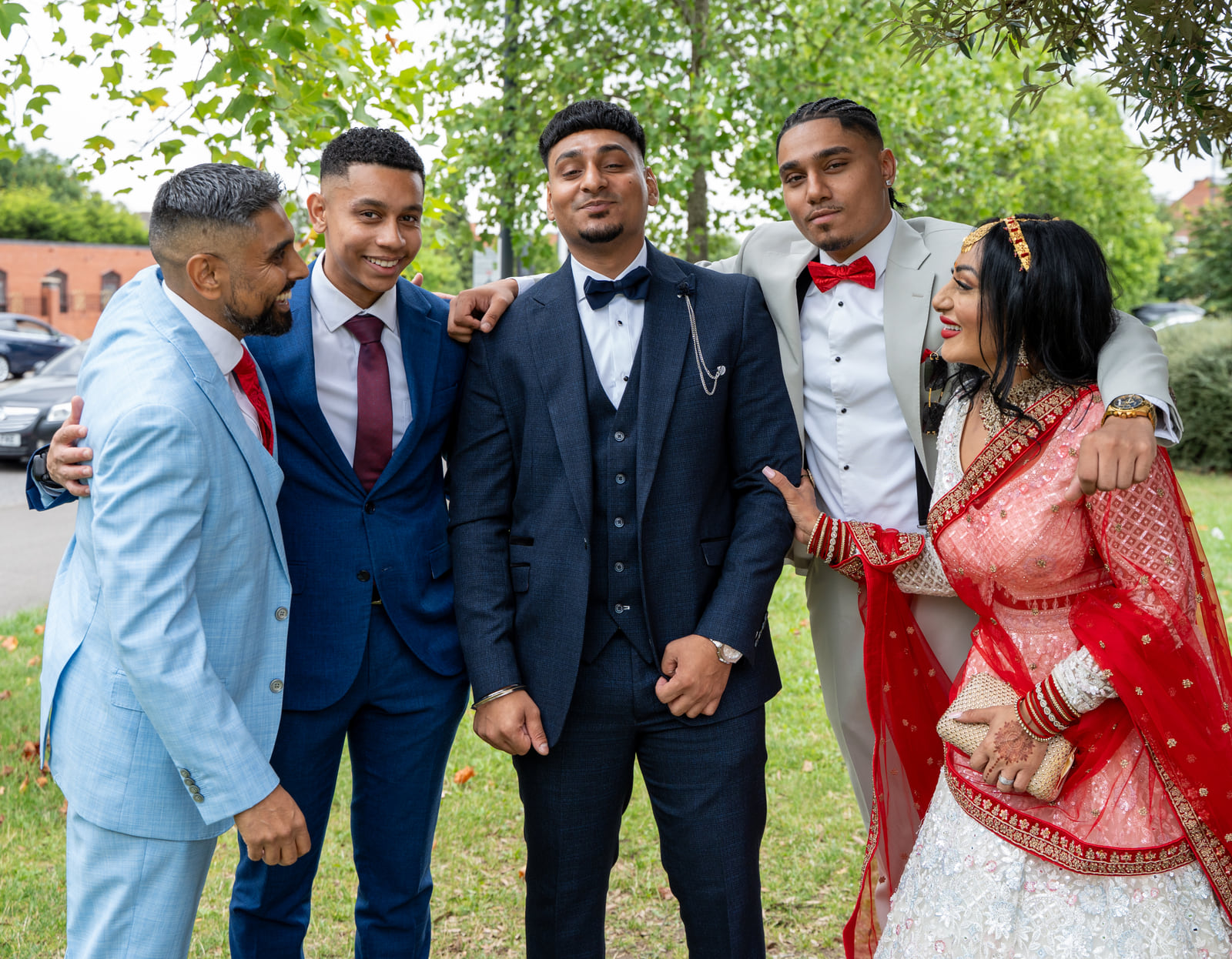 Reena Kaur in a red and white lehenga, arm in arm with four smiling men in suits