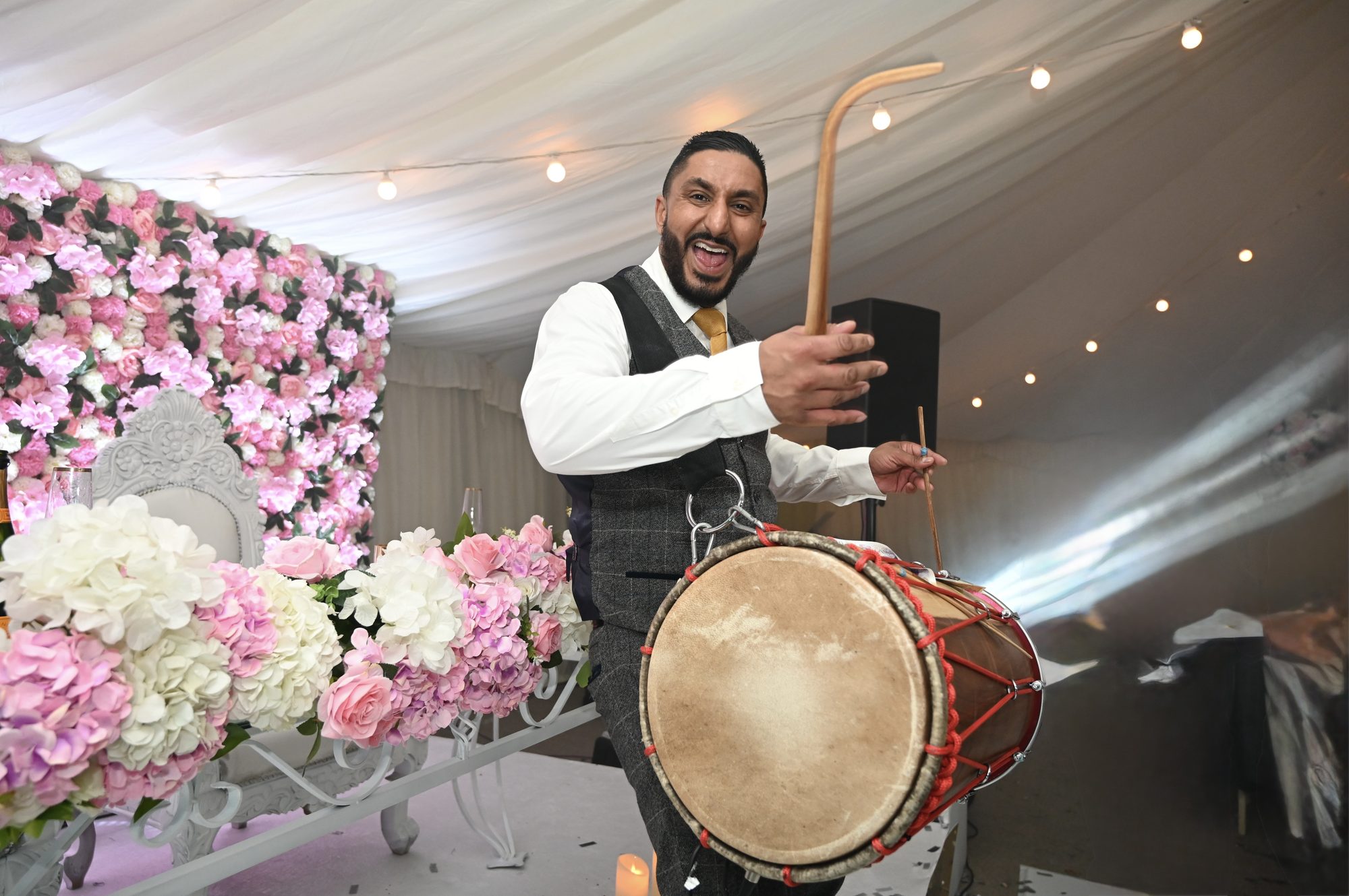 Dhol player performing at a marquee wedding beside a pink and white flower wall