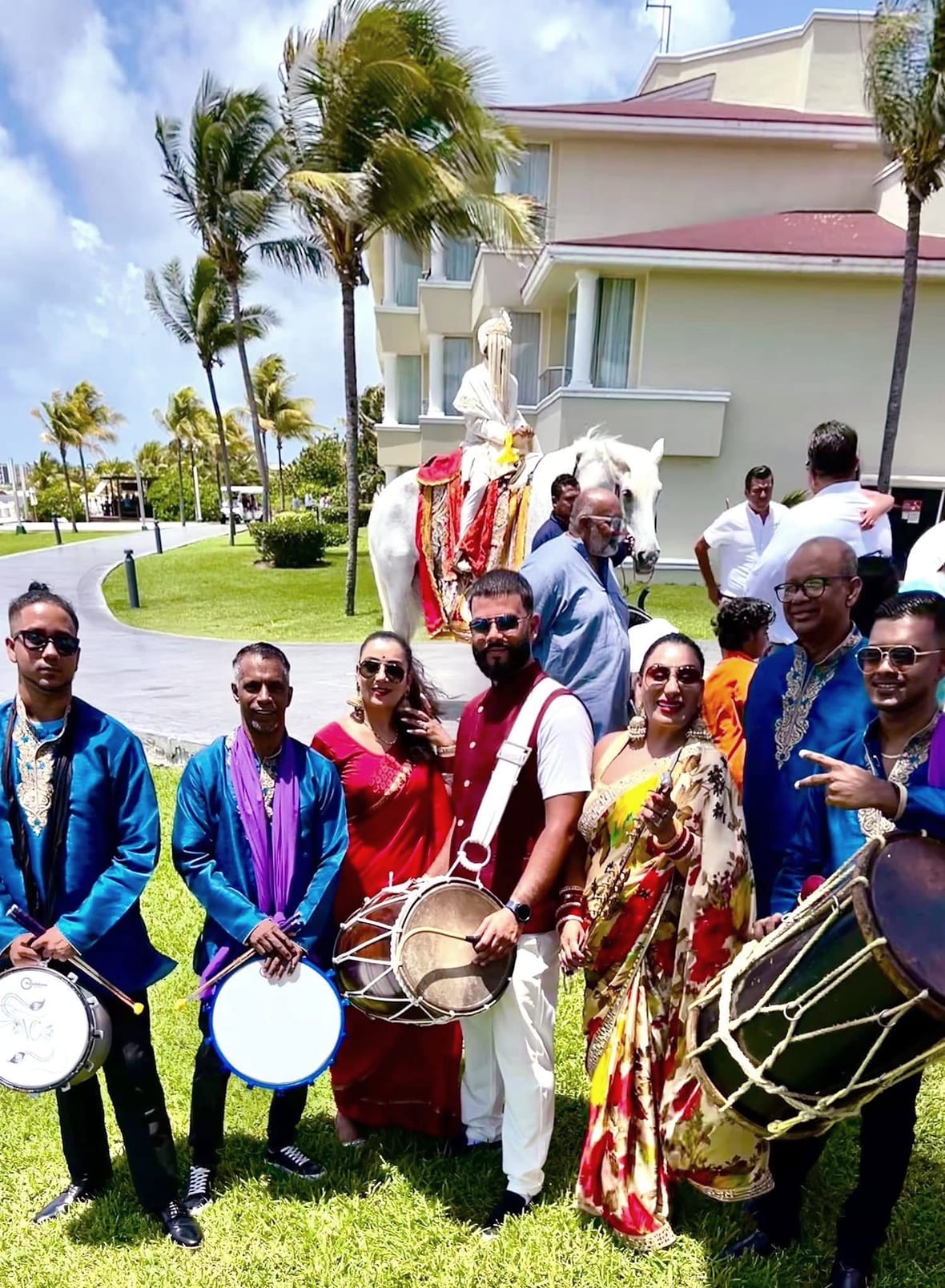 Dhol players in blue kurtas with a baraat party on a hotel lawn lined with palm trees, the groom on a decorated white horse behind them